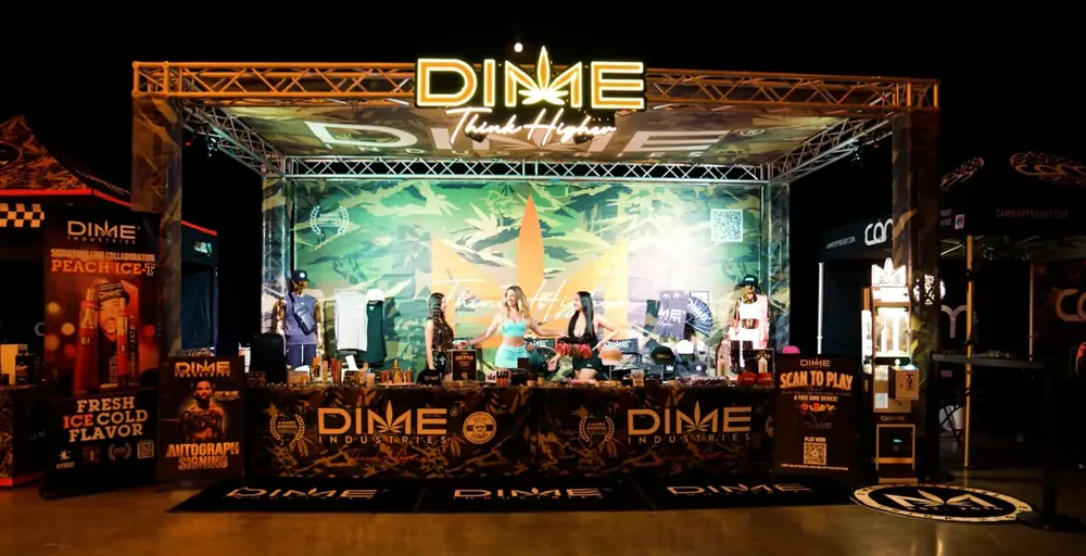 Dime Industries event booth under a lit truss with the Think Higher sign, camo backdrop, merch table and promo banners